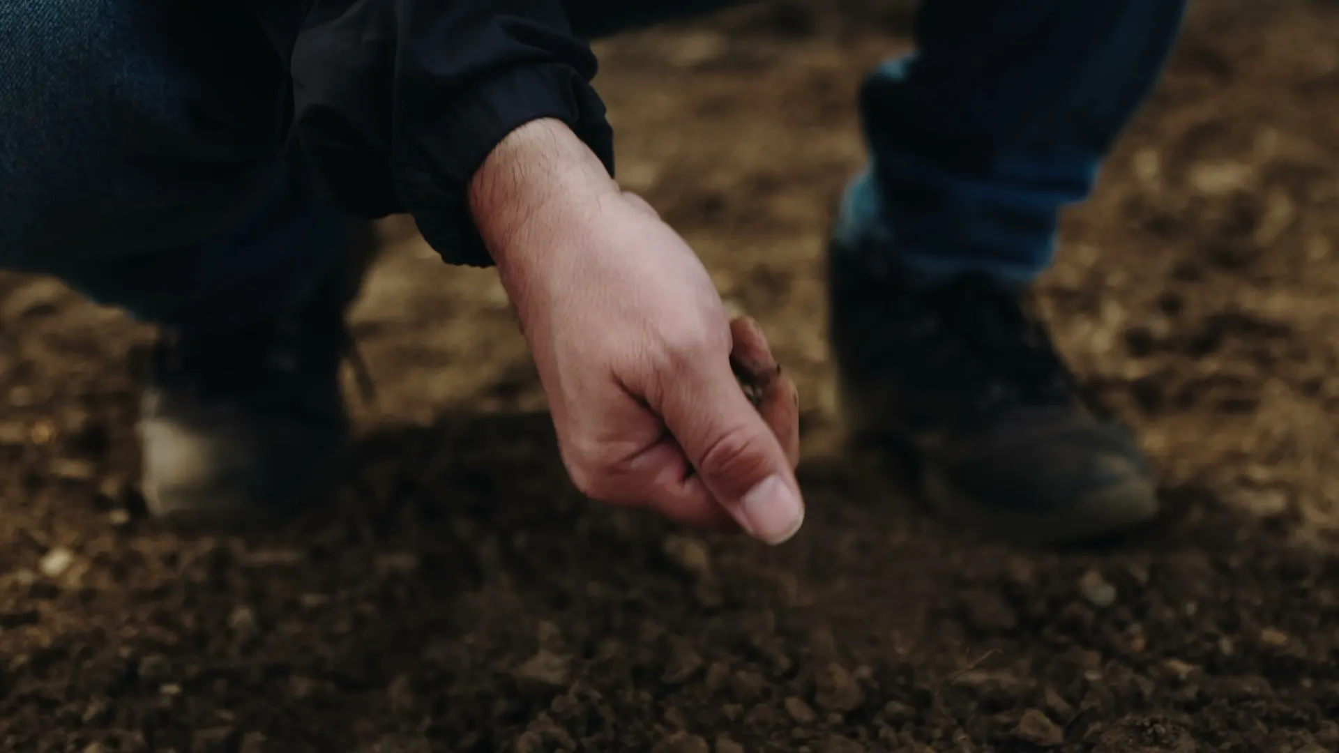 A hand letting a pinch of dark soil fall over freshly turned ground, boots behind it.