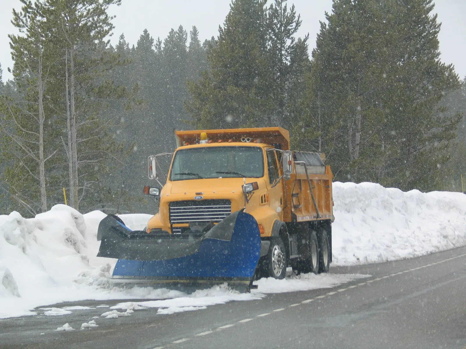 An orange plow truck pushing a bank of fresh snow along a forest road while it snows